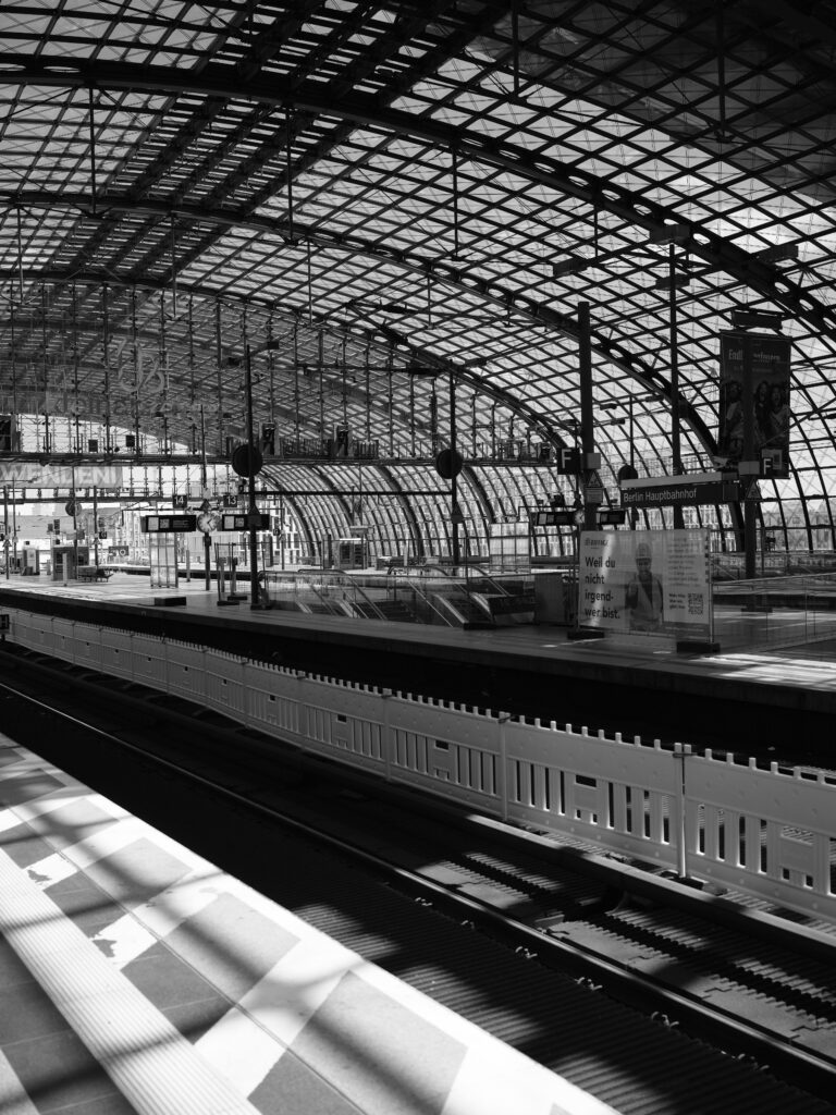 Sort-hvid fotografi af en stationshal med stor buet glas- og stålkonstruktion, tomme perroner, skiltning og spor i forgrund; stærke skygger og grafiske mønstre fra tagkonstruktionen.