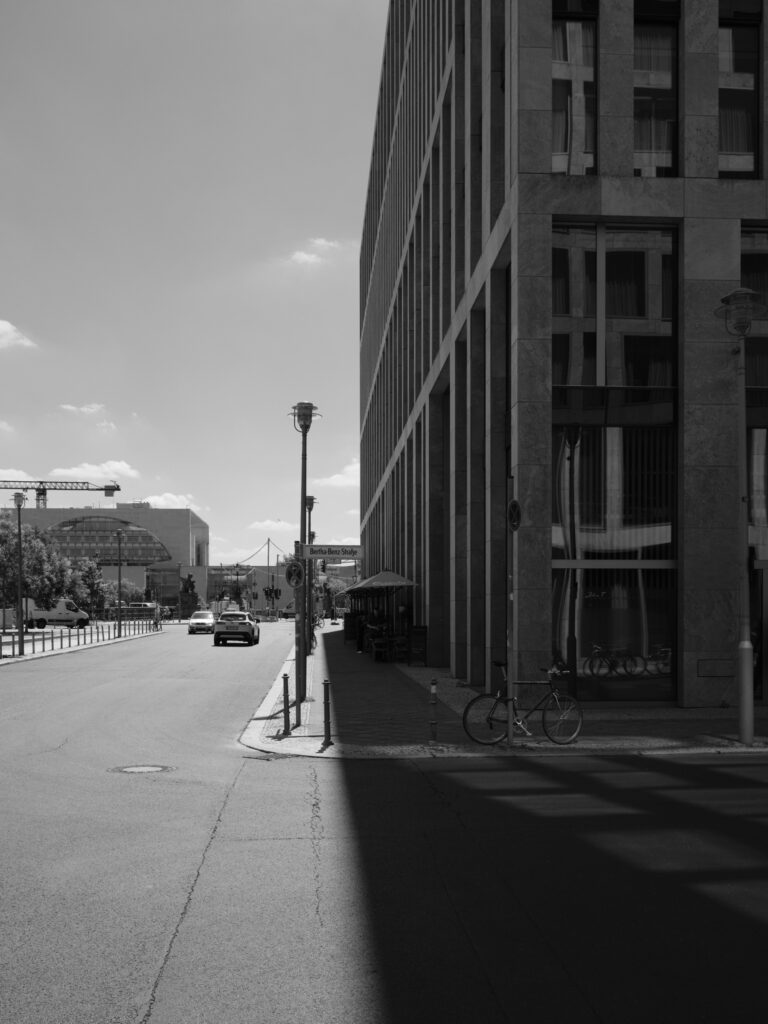 Sort-hvid fotografi af en bygade i Berlin med et højt kontorbyggeri til højre, en skarp skygge deler gaden i lys og mørke; et cykel parkeret ved fortovet, biler i baggrunden og en åben fornemmelse af perspektiv mod horisonten.