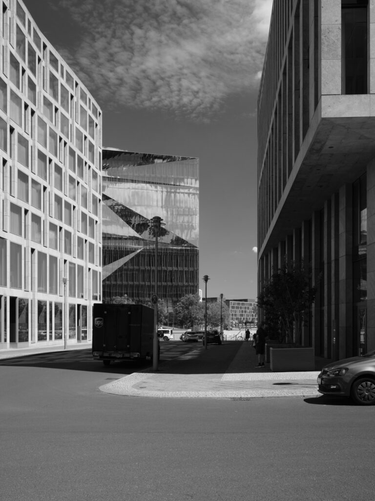 Sort-hvid fotografi af en byscene mellem moderne kontorbygninger. Billedet viser gadeareal i forgrund, en UPS-varebil i midten, og reflekterende glasfacade samt geometriske bygningsformer. Skyer ses i himlen over bygningerne.