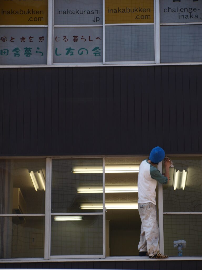 Fotografi af en person i blå hue og hvid/green trøje der arbejder ved en vinduesramme i en bygning med store vinduer; øvre del viser skilte med japansk tekst, indvendigt fluorescerende lys reflekteres i glasset.
